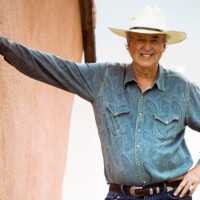 Tom Tatum Leaning Against Adobe Wall With Brush In Background - Featured Image